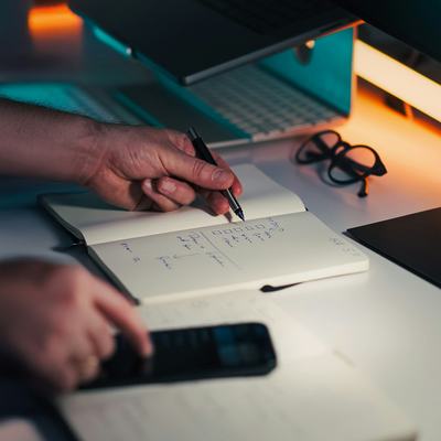 Manos anotando tareas en un cuaderno junto al portátil y el móvil, en un escritorio con luz cálida